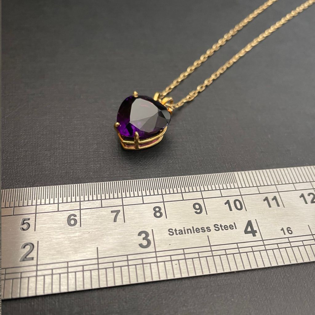 Heart-shaped amethyst pendant on a gold chain next to a metal ruler.