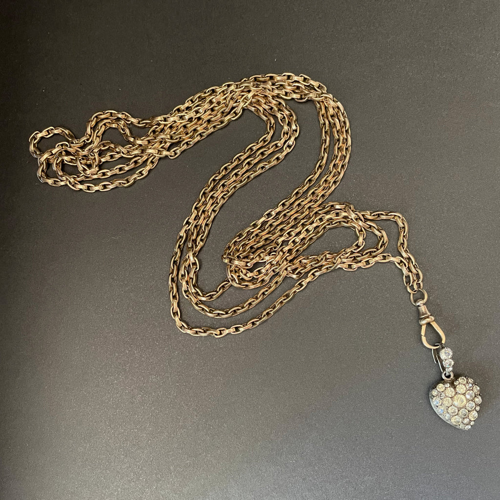 Gold long guard chain with swivel clasp holding a heart shaped pendant with colourless stones.