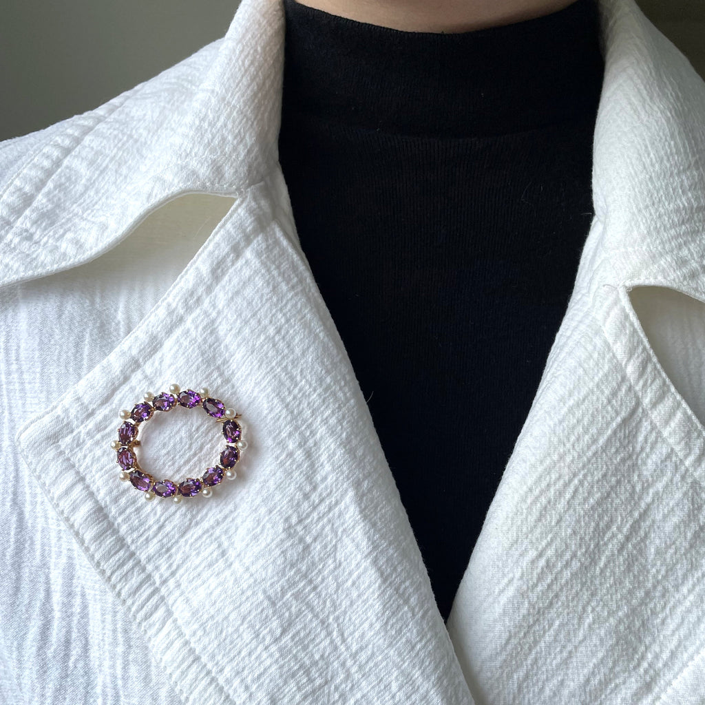 Woman wearing a white jacket with a black top and an oval amethyst brooch with pearls.