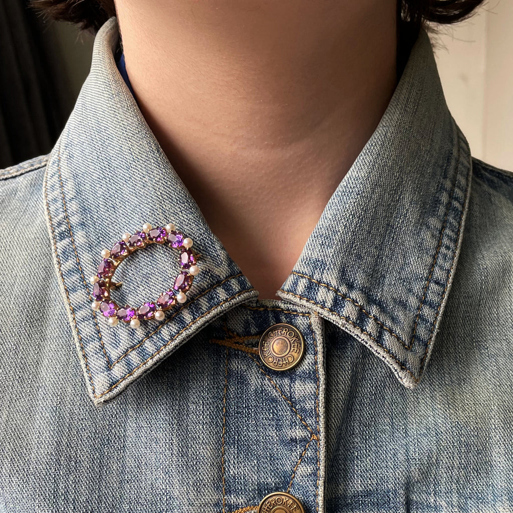 Oval gold brooch with amethysts and pearls in gold on a denim jacket collar.
